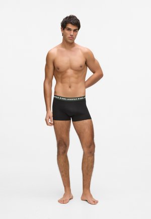 Kale man die zwarte Karl Lagerfeld boxershort draagt, blootsvoets staand tegen een effen witte achtergrond.