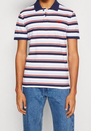 Homme portant un polo à manches courtes avec des rayures horizontales bleu marine, rouge, bleu clair et blanc, et un jean bleu, debout devant un fond uni.