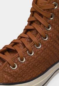 Κοντινό πλάνο καφέ πλεκτού ψηλού αθλητικού παπουτσιού με κορδόνια ίδιου χρώματος περασμένα μέσα από ασημένιες μεταλλικές οπές και λευκή λαστιχένια σόλα.