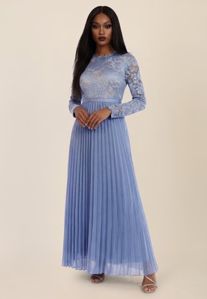 Frau trägt ein langes, plissiertes Lavendelkleid mit Spitzenmieder und Ärmeln mit Blumenmuster, steht mit gefalteten Händen vor neutralem Hintergrund.