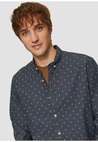 Navy-button-up shirt met kleine witte geometrische patronen, voorzien van een puntige kraag en een zachte stofstructuur, gedragen over een bruine T-shirt.