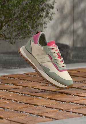 Beige, grøn og pink sneaker med tyk sål, svævende over trægulv med fliser, sløret udendørs baggrund med træ og sten.