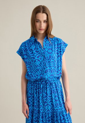 Abito camicia blu a fantasia con maniche corte, caratterizzato da una chiusura con bottoni e una vita regolabile con cordoncino. Il tessuto ha una superficie lucida e disegni intricati.