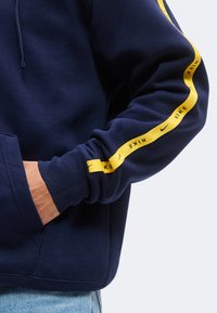 Μπλε σκούρο hoodie με κίτρινη ρίγα στο μανίκι με επαναλαμβανόμενα λογότυπα Nike, φορεμένο από ένα άτομο με ένα χέρι στην μπροστινή τσέπη.
