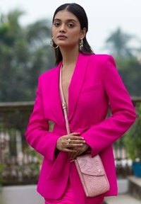 Fuchsia blazer met lange mouwen en een riem, gecombineerd met een lichtroze gestempelde crossbody tas. Model draagt hoepelringen en ringen.