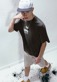 Ung mand iført en sort graphic t-shirt, beige shorts, hvid kasket og gule sko, stående på et hvidt gulv med sorte maleriplekker.
