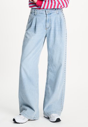 Persoon draagt lichtblauwe wijde jeans met studs aan de zijkant, witte sneakers en een rood-roze gestreept longsleeve top met gekruiste armen.