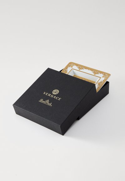 Čierna obdĺžniková krabička so zlatým textom, čiastočne otvorená, odhaľujúca biely tanier s ozdobným zlatým lemovaním a vzorom.