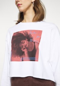 Sweatshirt cropped branco com uma estampa gráfica proeminente num fundo vermelho e texto. O material parece suave com um design casual.