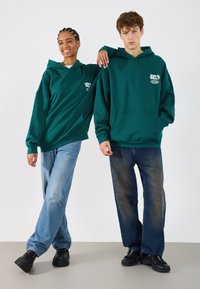 Grüne Kapuzenpullover mit Kängurutaschen, getragen über hellen und dunklen Jeans. Lässiger Schnitt; einer hat einen Logo-Aufdruck, während der andere schlicht ist.