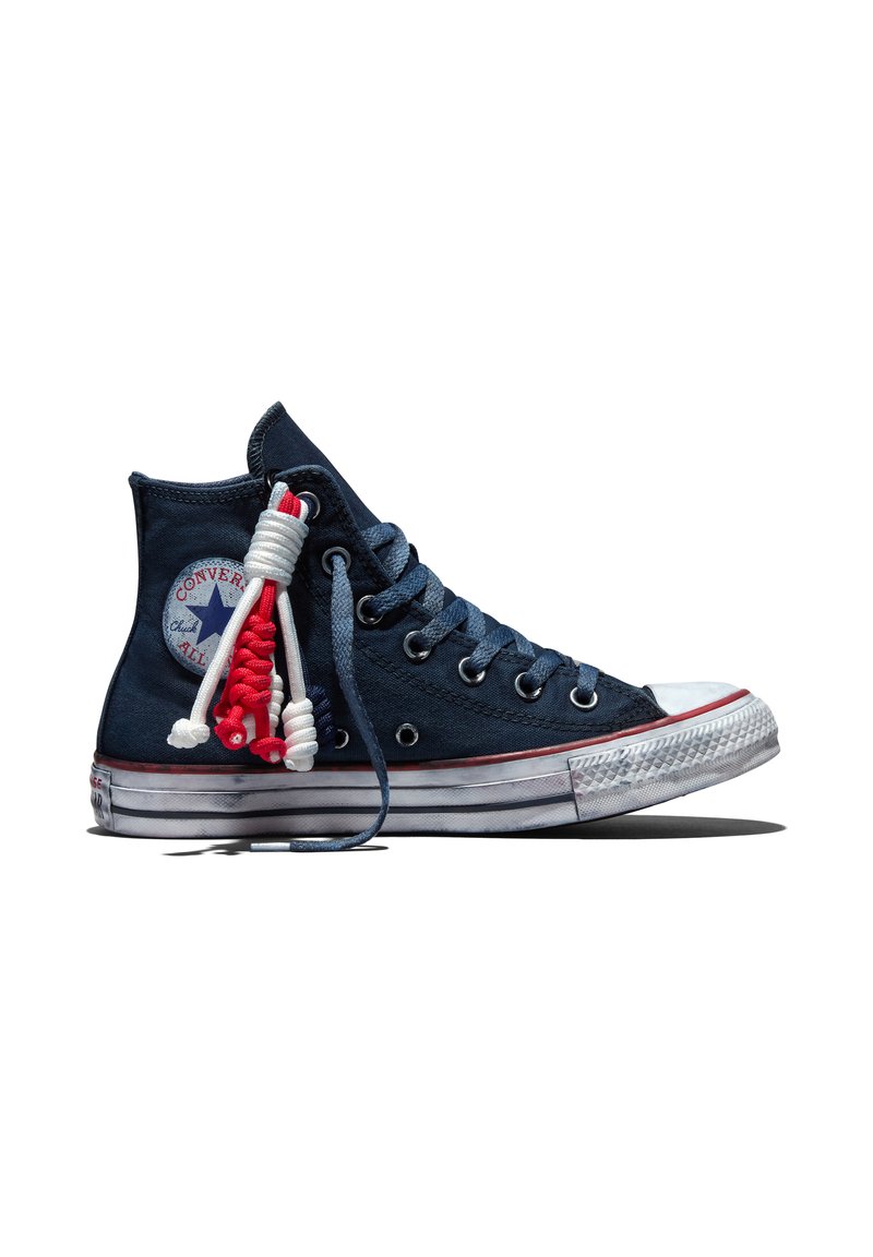 Navyblaue hohe Converse-Sneaker mit weißer Gummisohle, rotem Streifen und rot-weißen verknoteten Schnürsenkeln, die von der Seite hängen.