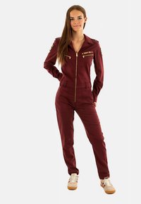 Bordeauxroter Jumpsuit mit Reißverschluss vorne, zwei Brusttaschen und goldenen Akzenten. Mit langen Ärmeln und zulaufenden Beinen, kombiniert mit beige-roten Turnschuhen.
