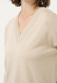 Pull beige à col en V en tissu doux. Présente un col côtelé et une texture lisse. Coupe ajustée qui met en valeur le cou et les épaules.