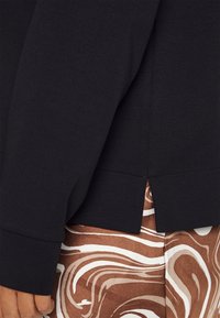 Sudadera negra de manga larga con una abertura lateral. El material parece suave y tiene una textura lisa. Combinada con pantalones de patrón mármol en tonos marrón y blanco.