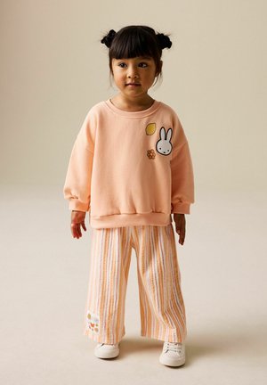 Young child with dark hair in buns wearing a peach sweatshirt with bunny and flower patches, striped wide pants, and white sneakers, standing.