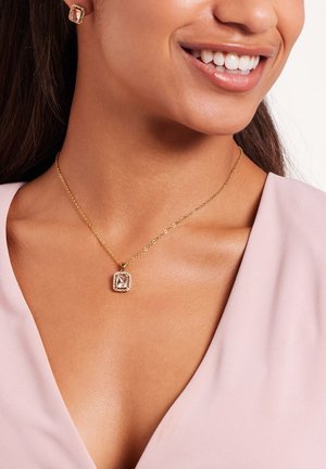 Collier en or avec un pendentif carré présentant une pierre centrale et des accents environnants, porté sur un haut rose clair.