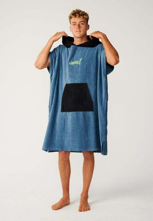 Joven de pie descalzo vestido con un poncho toalla azul extragrande con capucha, un bolsillo frontal negro y un logo amarillo.