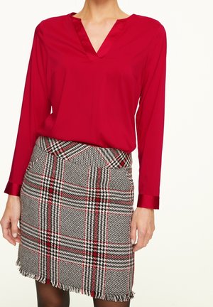 Woman wearing a red long-sleeve blouse tucked into a black, white, and red plaid skirt with fringed hem, standing against a plain background.