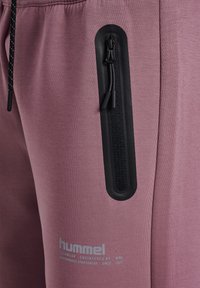 Ljuslila joggers med en svart dragkedjeficka, dragsko i midjan och logodetalj i silver. Slät tygtextur med kontrasterande detaljer.
