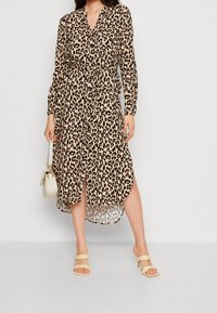 Robe à manches longues imprimée léopard avec une taille cintrée. Elle présente un devant boutonné et un ourlet asymétrique. Portée avec des sandales neutres et un sac à main.