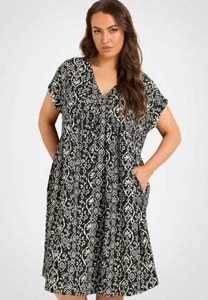 Kvinde iført en knælængde kjole med sort og hvidt mønster, korte ærmer, V-hals og lommer, stående mod en enkel lys baggrund.