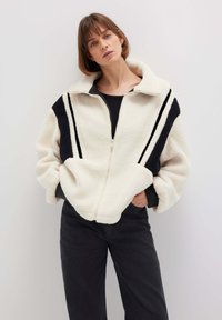 Fleece-Zip-Jacke in Creme mit schwarzen Akzenten und Streifen. Verfügt über einen hohen Kragen und Fronttaschen. Texturiertes Material mit weicher Oberfläche.
