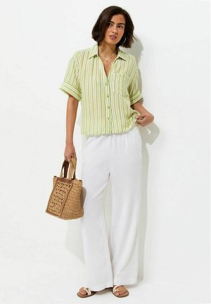 Mujer vestida con una camisa de rayas verde y blanca abotonada, pantalones blancos de pierna ancha, sandalias y sosteniendo un bolso tejido marrón.