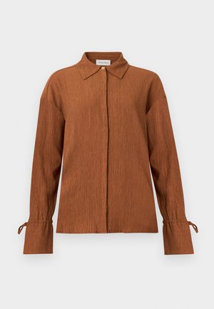 Καφέ μπλούζα με μακριά μανίκια και υφή, με μυτερό γιακά, κρυφά κουμπιά στο μπροστινό μέρος και δετούς μανσέτες σε μονόχρωμο φόντο.