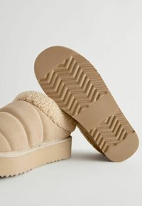 Chausson beige avec intérieur doux en fourrure ; extérieur matelassé ; semelle en caoutchouc avec motif de silhouette en zigzag ; design à profil bas.