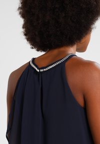 Top sans manches bleu marine avec un dos en forme de clé et un détail de col argenté embellit, fabriqué dans un tissu doux et fluide.