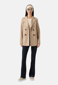 Beige dobbeltradet blazer med sorte knapper, parret med mørke denim flare jeans og cremede sneakers. Tilbehøret inkluderer hvide solbriller og et pandebånd.