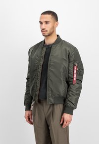 Olive green bomber jacket in shiny nylon with ribbed cuffs and hem. Features an angled zip pocket with a red tab on the sleeve.