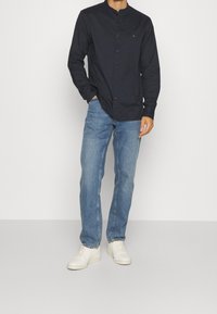 Koszula zapinana na guziki w granatowym kolorze z bawełny, kołnierzyk stójkowy, długie rękawy, z przodu zapięcie na guziki, w połączeniu z jasnoniebieskimi dżinsami i białymi trampkami.