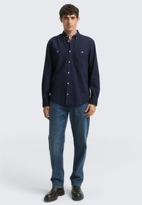 Navyblaues Hemd mit strukturiertem Design, zwei Brusttaschen und weißen Knöpfen, kombiniert mit blauen Jeans und schwarzen Stiefeln.