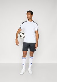 Chemise de sport blanche avec des rayures noires, short gris, chaussettes de football blanches avec des rayures bleues et chaussures de football blanches. Tiens un ballon de football noir et blanc.