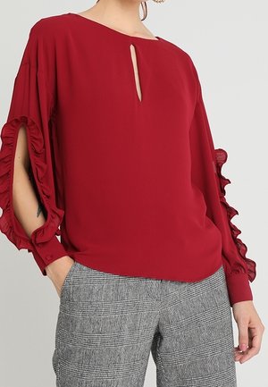Rode blouse met keyhole-hals en gerimpelde open mouwen gecombineerd met grijze geruite broek bij een staand persoon.
