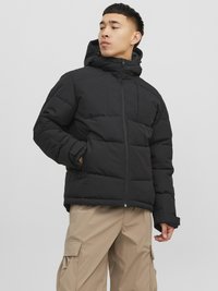 Giubbotto bomber nero con cappuccio, caratterizzato da sezioni trapuntate e una zip frontale. Abbinato a pantaloni cargo beige chiaro.