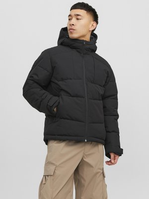 Giubbotto bomber nero con cappuccio, caratterizzato da sezioni trapuntate e una zip frontale. Abbinato a pantaloni cargo beige chiaro.