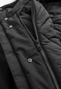 Czarne wnętrze kurtki puchowej z pikowaną fakturą, srebrnymi zatrzaskami i czarnym zamkiem błyskawicznym, o gładkim, matowym wykończeniu.