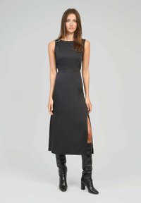 Robe maxi noire sans manches avec une fente latérale, mettant en valeur un tissu lisse et un accent métallique sur l'épaule. Portée avec des bottes noires jusqu'au genou.