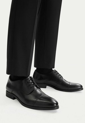 Schwarze, aus Leder gefertigte Halbschuhe mit Kappe, getragen zu schwarzer Hose und schwarzen Socken auf hellem Hintergrund.