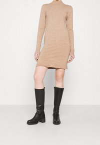 Robe longue à manches longues en maille beige avec un col rond côtelé et des poignets assortis, associée à de grandes bottes en cuir noir à talon carré et fermeture éclair.
