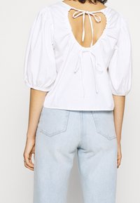 Bílá bavlněná blůza s hlubokým otevřeným zadním výstřihem, svázaná odpovídajícím materiálem, a nabíranými rukávy. Spodní lem je rovný a zkrácený.