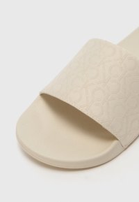 Beige Rutschsandal mit Obermaterial aus strukturiertem Stoff, das ein dezentes Logomuster aufweist, runder Zehenbereich und geformte Gummisohle.