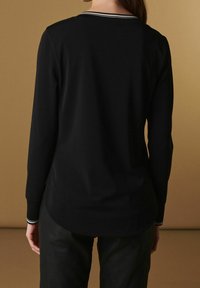 La maglietta nera a maniche lunghe presenta un colletto a girocollo con una striscia bianca. Realizzata in tessuto morbido, ha una vestibilità rilassata e una texture liscia.