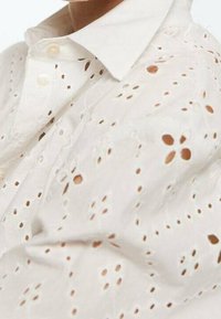 Bílá bavlněná košile s laserovým řezáním květinových vzorů, špičatým límečkem a knoflíkovým zapínáním. Měkká textura s lehkým vzhledem.