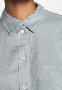 Camisa de lino gris claro con un cuello clásico, siete botones en la parte delantera y un bolsillo en el lado izquierdo del pecho. Tela suave y texturizada.