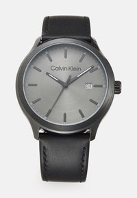Reloj de pulsera Calvin Klein con una esfera gris, marcadores de hora negros y una pantalla de fecha. Presenta una correa de cuero negro y una caja redonda lisa.