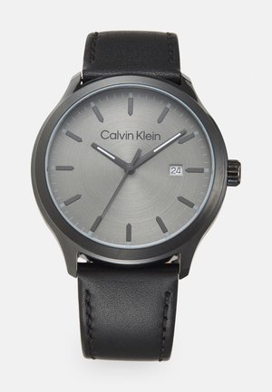 Montre-bracelet Calvin Klein avec un cadran gris, des index noirs, et un affichage de la date. Dispose d'un bracelet en cuir noir et d'un boîtier rond lisse.