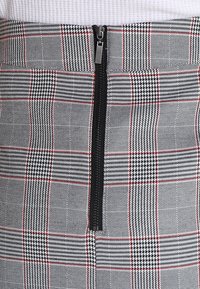 Gonna scozzese grigia con accenti rossi, caratterizzata da una cerniera nera al centro. Il materiale ha una consistenza liscia con un design strutturato.